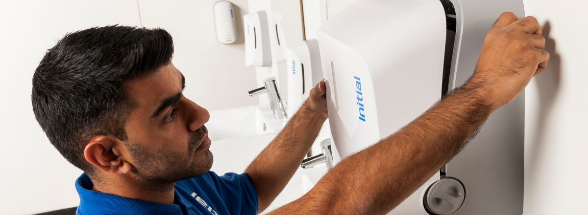 A technician wearing a blue polo shirt installs or services a white wall-mounted hygiene dispenser branded with the "Initial" company logo in a clean restroom setting.